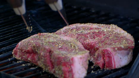 un par de pinzas para carne juegan con un filete de costilla crudo en una parrilla y lo presionan junto con otro filete