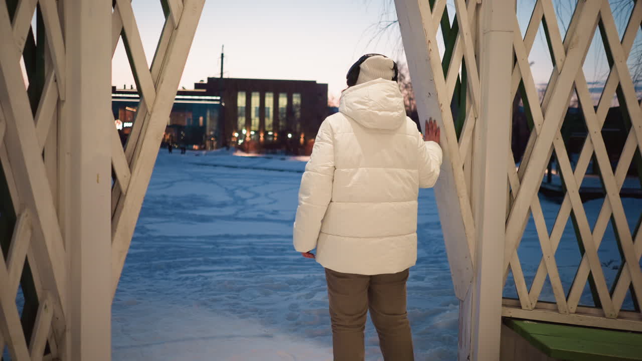 Back view of woman wearing puffer coat and beanie, gently swaying inside decorated pavilion lit by string lights in snowy park under evening sky, hands on lattice as soft breeze hints motion