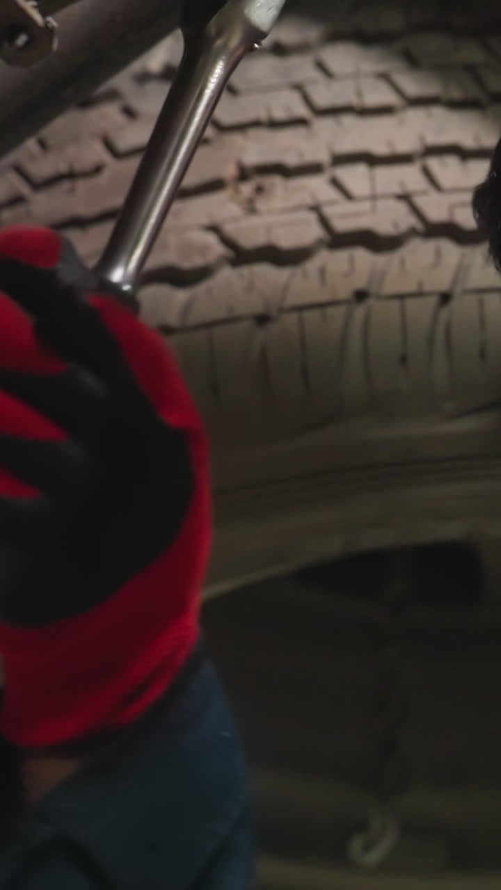Indian engineer working under car, tightening bolt with focused expression, light reflecting on face, wearing gloves and blue uniform, engaged in automotive repair, inspecting vehicle mechanics