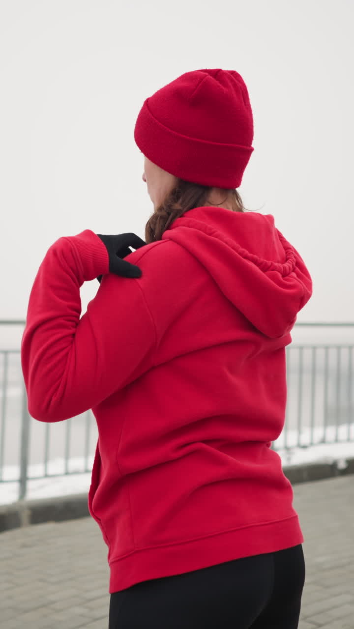 back view of woman in red hoodie and beanie outdoors performing arm stretches by lifting hands to shoulders and turning arms, background features misty atmosphere with river and iron railing