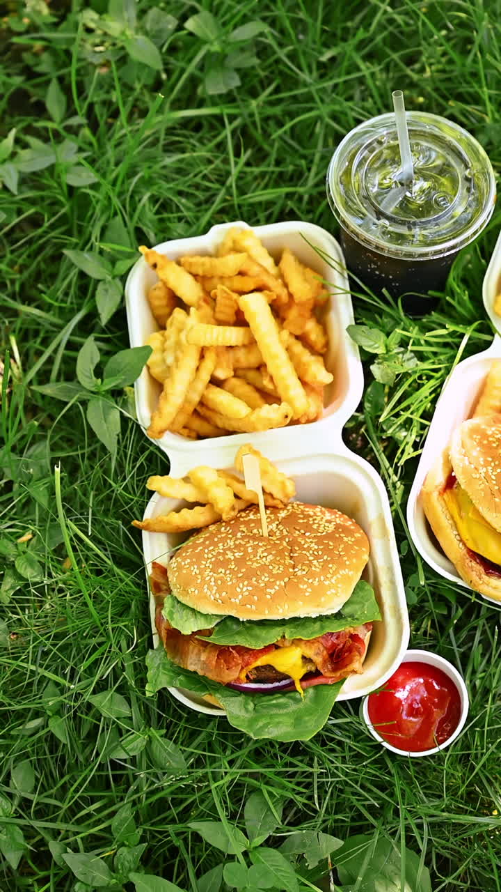 Hambúrguer e batatas fritas em embalagem para viagem com ketchup ao ar livre. Hambúrguer delicioso com alface e bacon servido com batatas fritas e ketchup em embalagem de papel na grama em um piquenique