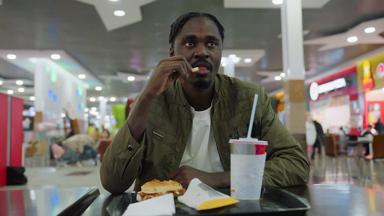 Focused young man wearing green jacket chewing french fries in vibrant food court setting, seated at table with burger, fries, paper cup on tray, bright lights and blurred shoppers in background