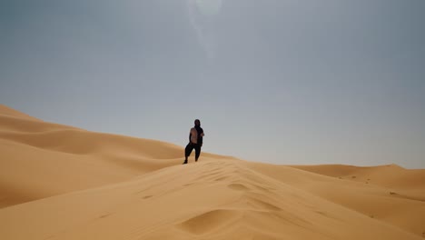Person stands on the crest of a golden sand dune under a clear sky in Merzouga, Sahara Desert, Morocco