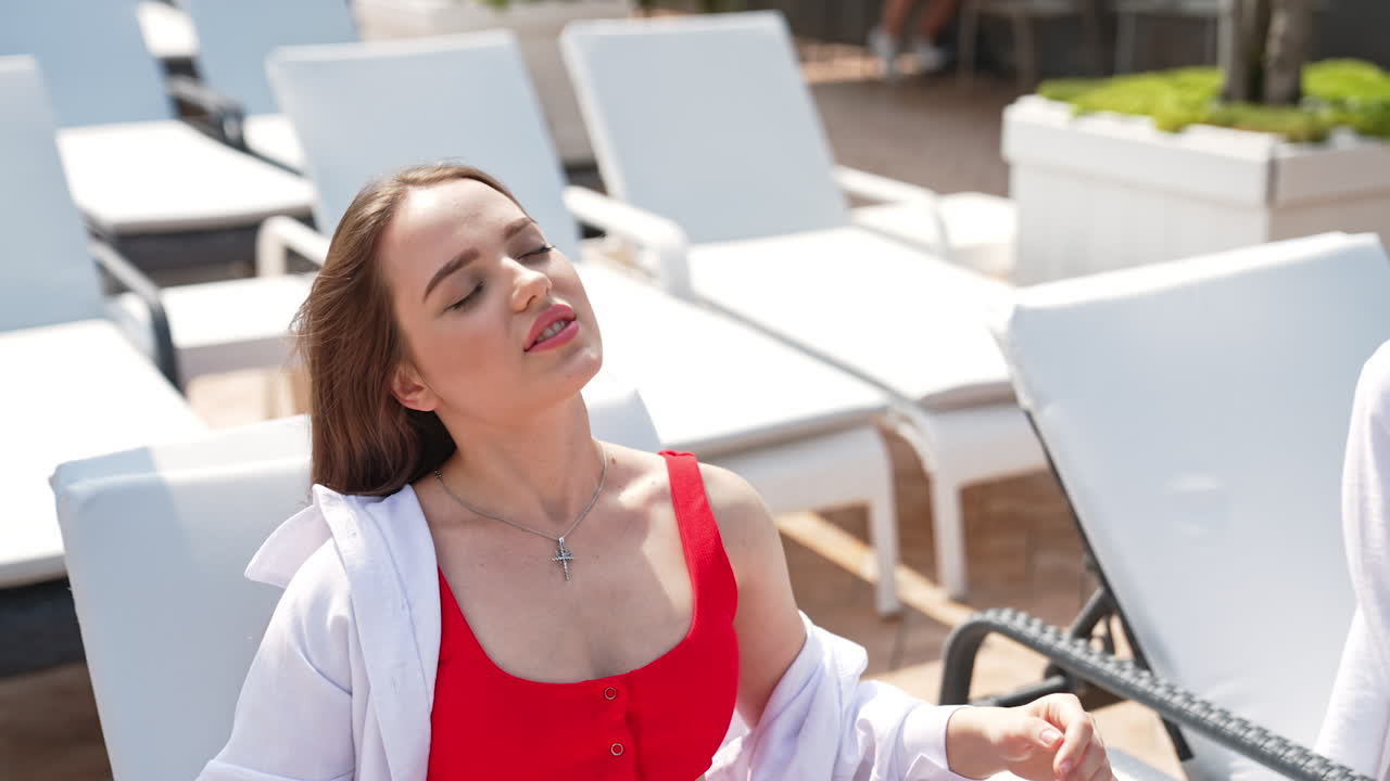 Young sexy lady in red swimsuit and white shirt enjoying sun. Girl lies in white deckchair looking into camera.