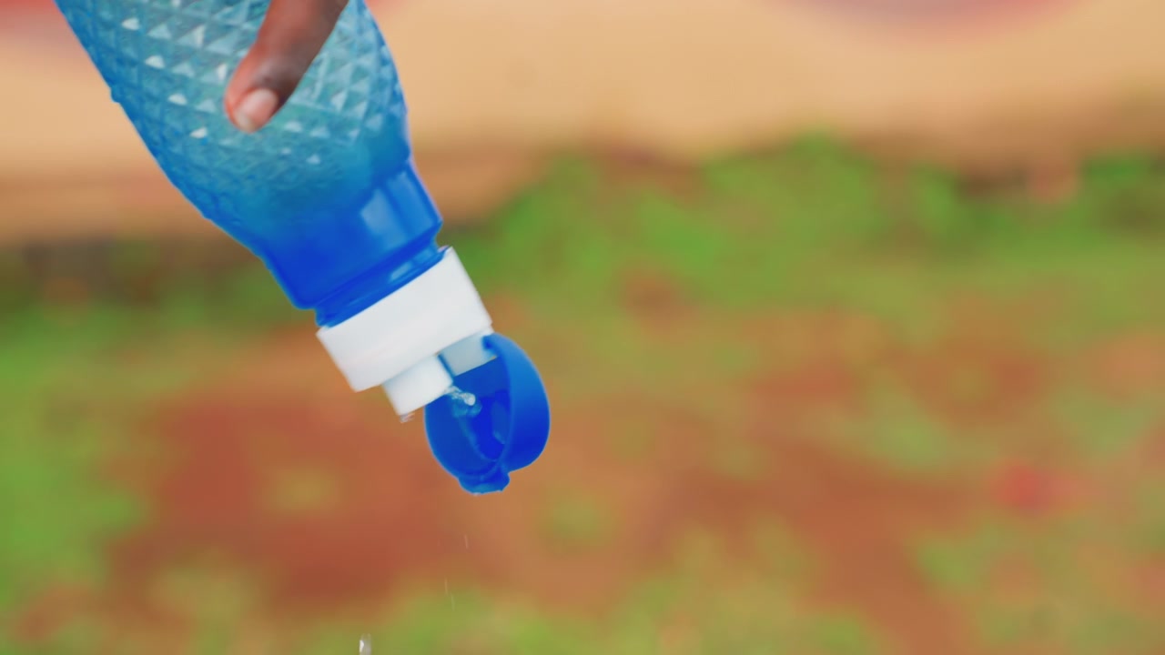 Close-up van hand die water giet buiten, blauwe plastic fles gekanteld met druppels die op aarde en groene spruiten vallen, volwassen hand stabiel, geringe scherptediepte, warm zonlicht, zachte beweging suggererend