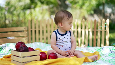 cute baby boy in sleeveless t-shirt sits on the plaid on the ground watching soap bubbles fly. adorable kid resting outdoors in summer.