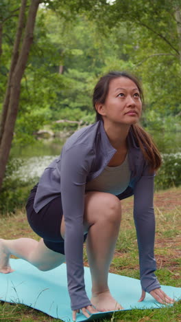 Video-Vertical-De-Una-Mujer-Vestida-Con-Ropa-Deportiva-Haciendo-Estiramientos-De-Yoga-Sobre-Una-Colchoneta-En-Un-Bosque-Junto-A-Un-Lago-O-Río-Disfrutando-De-La-Paz-Y-La-Belleza-De-La-Naturaleza-Filmado-En-Tiempo-Real-3