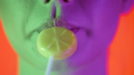 Primer-Plano-De-Labios-De-Una-Joven-Sexy-Mientras-Lame-Una-Dulce-Paleta-De-Limón-Con-Placer-Con-Su-Lengua-Sobre-Un-Fondo-Naranja-Con-Contraste-Verde-Púrpura-En-La-Cara-En-Cámara-Lenta