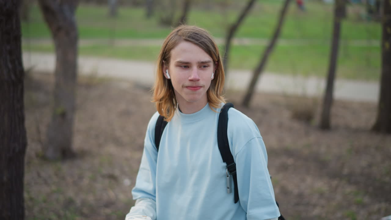 estudiante joven tomando café caliente cerca de un árbol, con auriculares, primer plano de perfil, expresión consciente, vaso desechable en las manos, sonrisa sutil, hierba de primavera, poca profundidad de campo, tonos cálidos