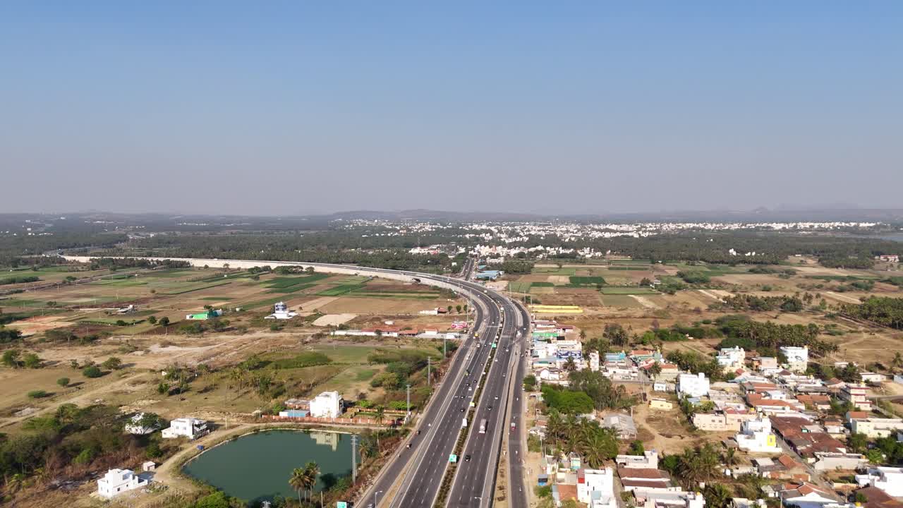 An aerial view of a major highway cutting through the Indian countryside on a sunny day. wide, multi-lane road with moderate traffic, surrounded by farmland, small residential areas, and vegetation