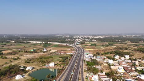 an aerial view of a major highway cutting through the indian countryside on a sunny day. wide, multi-lane road with moderate traffic, surrounded by farmland, small residential areas, and vegetation