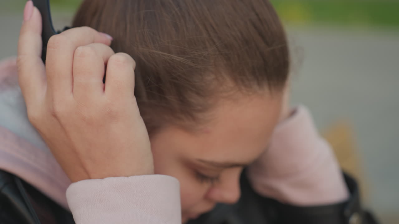 Close up of white woman with polished nails wearing pink hoodie and black leather jacket seated at wooden table slowly lifts headset with wind gently blowing hair with blur background