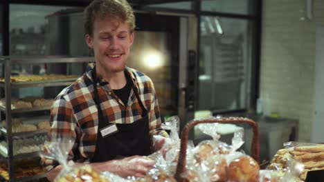 positivo, sonriente empleado de la tienda en delantal negro poniendo pastelería fresca, muffins en la vitrina de pie en la hermosa tienda con productos de panadería. vista frontal