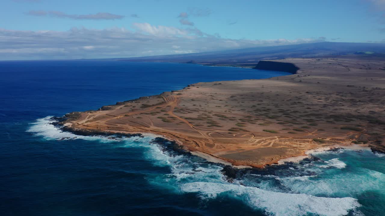 Foamy blue waves slam into dry rocky cliffs, revealing an expansive coastline of dusty trails and open sky stretching toward a distant horizon.