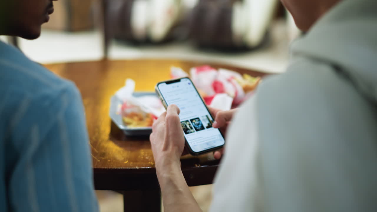 Back view of two friends leaning together browsing mobile phone in casual setting, shoulders visible, focus on screen interaction and companionship, one wearing blue shirt and another white
