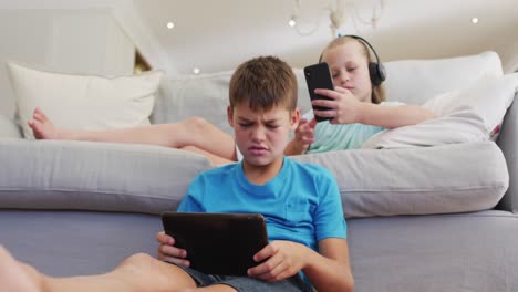 Portrait-of-caucasian-brother-and-sister-lying-on-sofa-using-tablet-and-smartphone