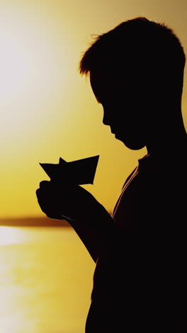 young boy is making a paper ship standing near the river at the evening sunset. silhouette of the boy playing with paper boat outside vertical video