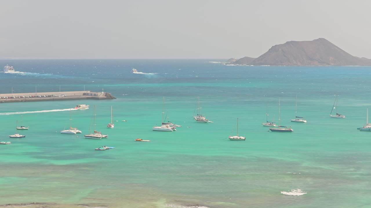 Static drone view over turquoise waters off Fuerteventura, showing anchored boats near a pier and the volcanic silhouette of Lobos Island rising in the distance under clear skies