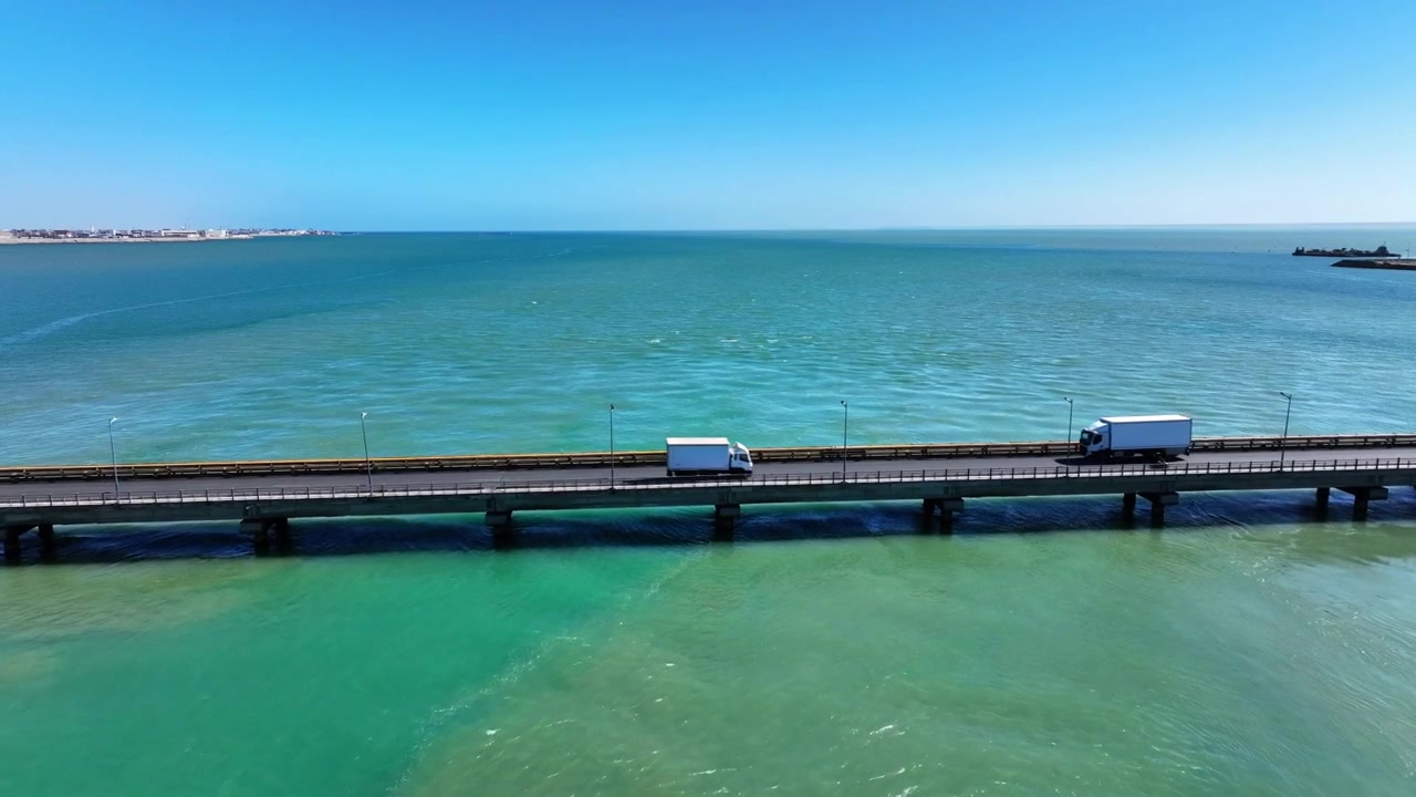 モロッコ南部、ダフラ湾のターコイズブルーの海をまたぐ海岸橋のドローン空撮。この地域で最も特徴的な景観の一つを横断し、沿岸地域を結ぶ近代的な海洋インフラです。
