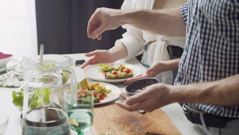 Primer-Plano-De-Un-Hombre-Que-Agrega-Una-Mezcla-De-Semillas-A-Una-Deliciosa-Ensalada-En-Su-Plato,-Mientras-Su-Esposa-Está-De-Pie-Junto-A-él-Con-Su-Plato