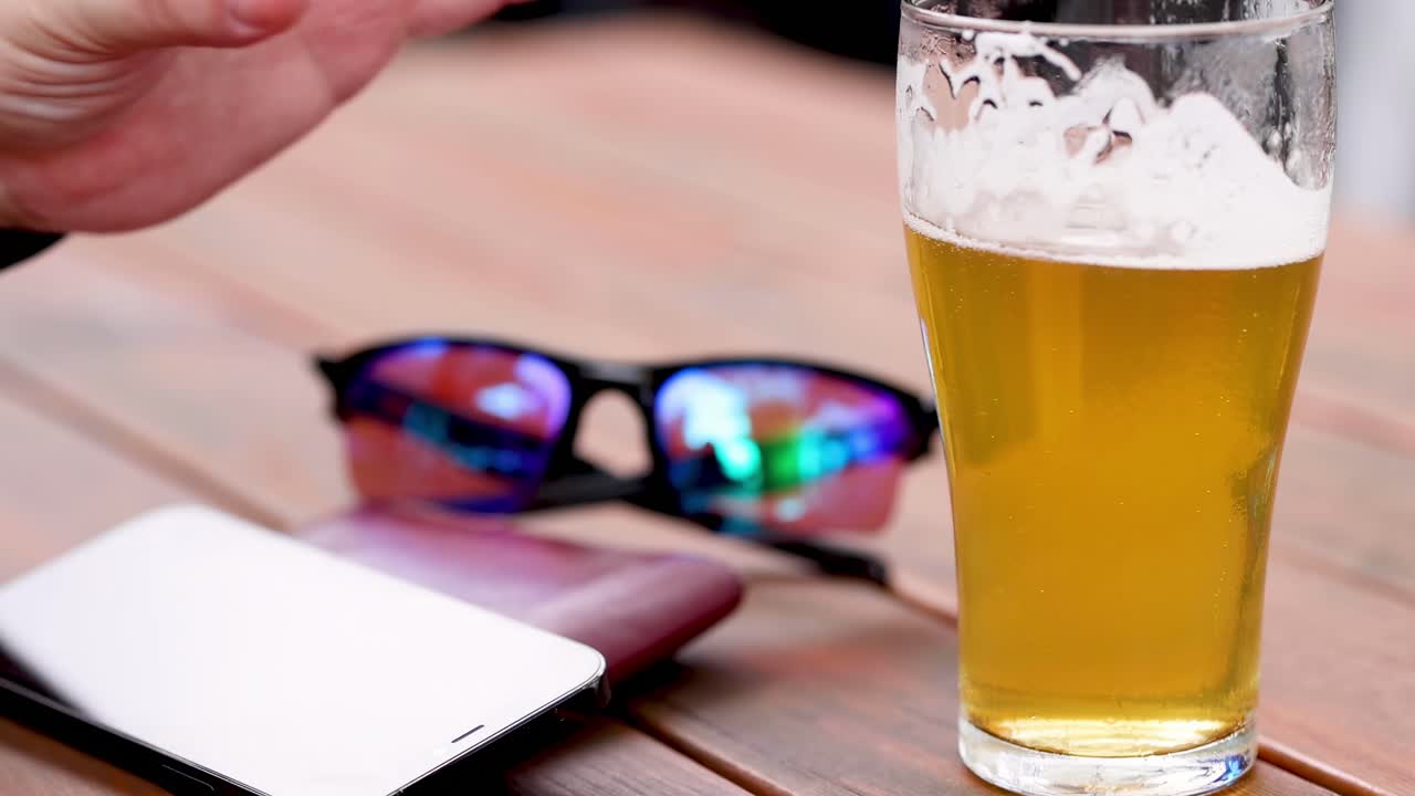 A person picks up a beer glass from a wooden table beside sunglasses and a smartphone.