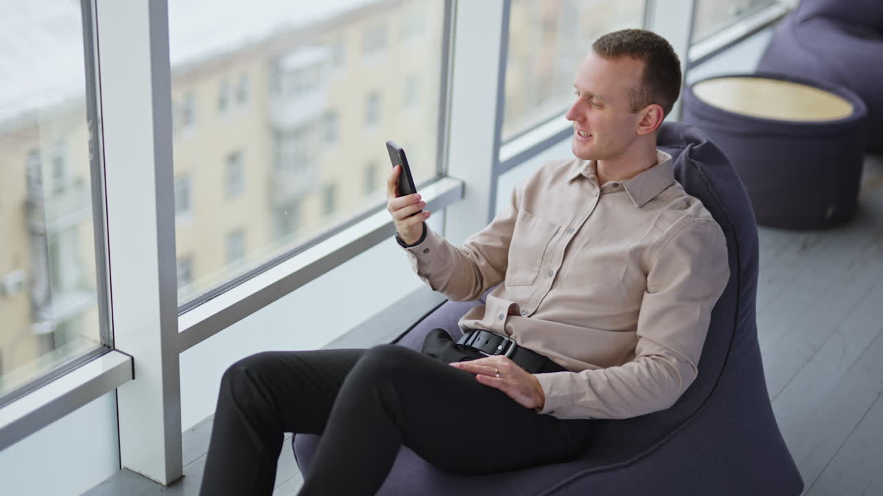 Relaxed young man sits in bean bag chair with phone in hand. Male having rest enjoying city panorama in big windows.