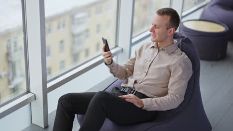 relaxed young man sits in bean bag chair with phone in hand. male having rest enjoying city panorama in big windows.