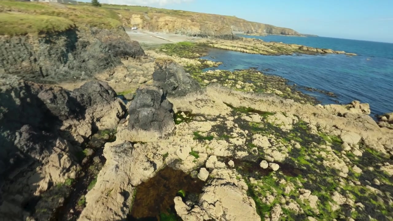 Un drone FPV sfreccia basso su distese di alghe verdi verso scogliere scure e le acque blu dell'Atlantico lungo la Costa del Rame in Irlanda