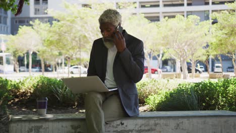 Afroamerikanischer-älterer-Mann,-Der-Auf-Dem-Smartphone-Spricht,-Während-Er-Im-Firmenpark-Einen-Laptop-Benutzt