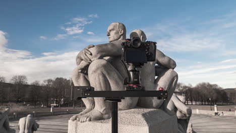 Cámara-Compacta-En-Un-Deslizador-Disparando-Una-Escultura-De-Granito-De-Dos-Hombres-Sentados-Espalda-Con-Espalda-Con-Los-Brazos-Cruzados-Sobre-Un-Pedestal-En-El-Parque-Frogner,-Oslo,-Noruega