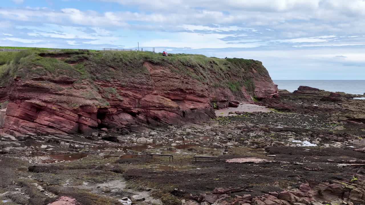 Camera pans right revealing rocky shore, red sandstone cliffs, seaweed, and overcast coastal landscape