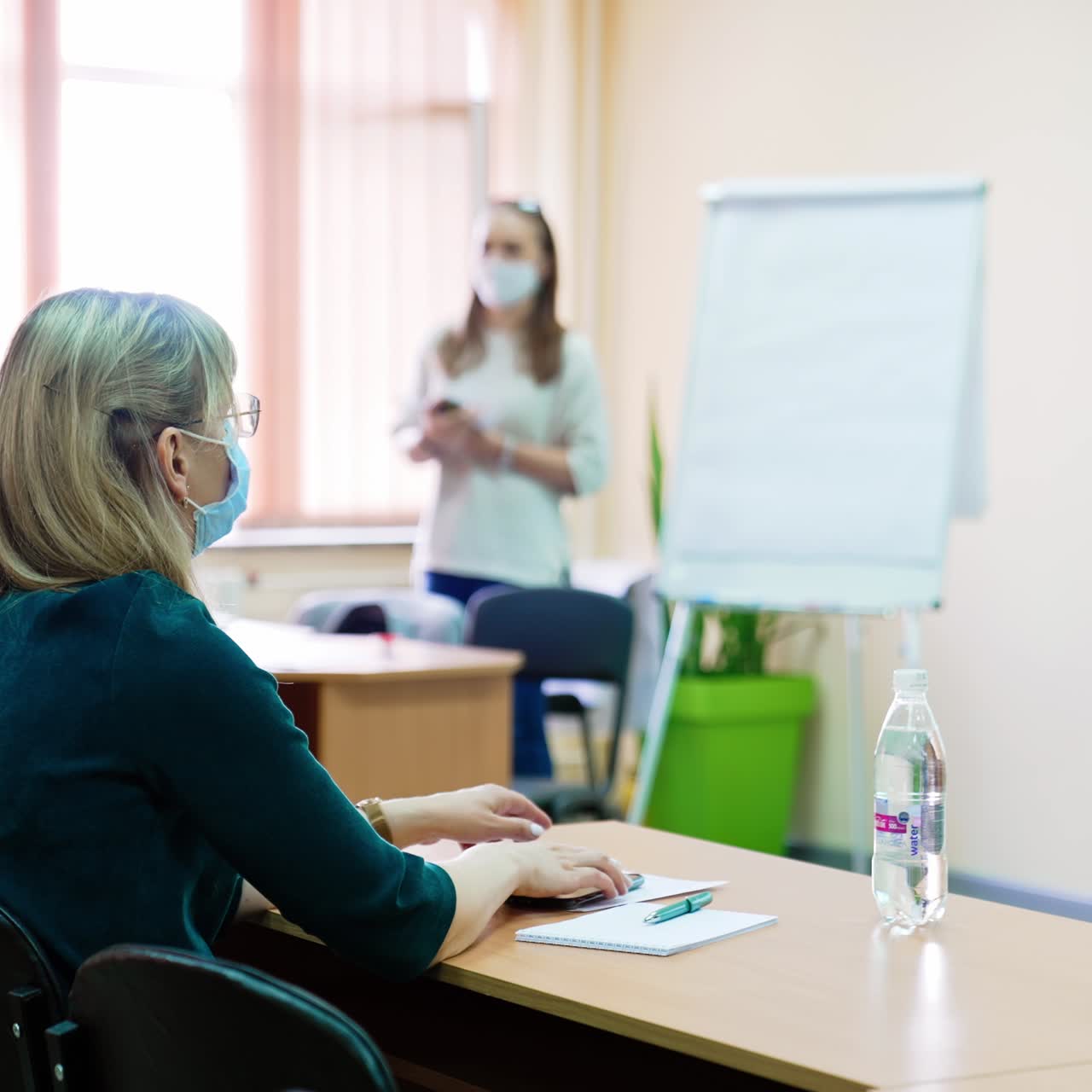 Estudiantes con mascarillas quirúrgicas tomando apuntes durante una clase. Mujeres sentadas en pupitres escuchando a la profesora. Fondo difuminado