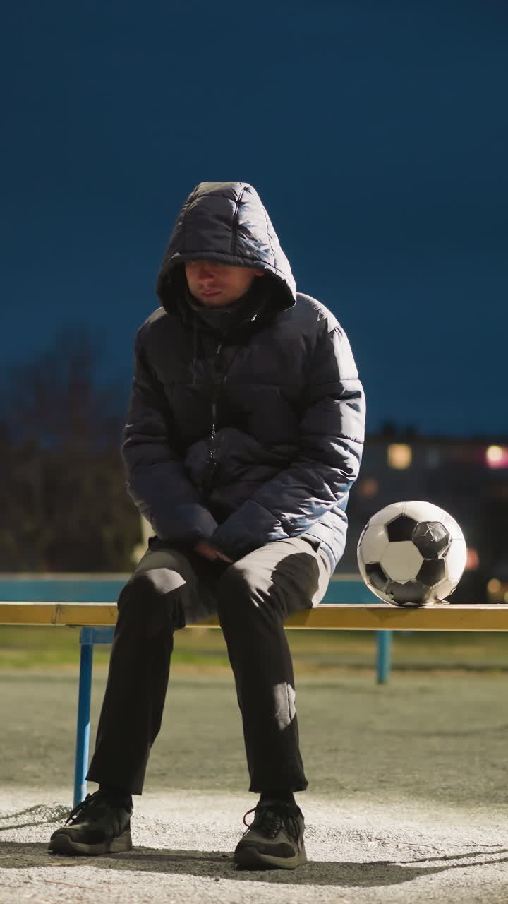 вдумчивый человек сидит в одиночестве на скамейке на открытом воздухе, с руками, сцепленными между ногами, рядом с ним находится футбольный мяч, на заднем плане кто-то едет на велосипеде