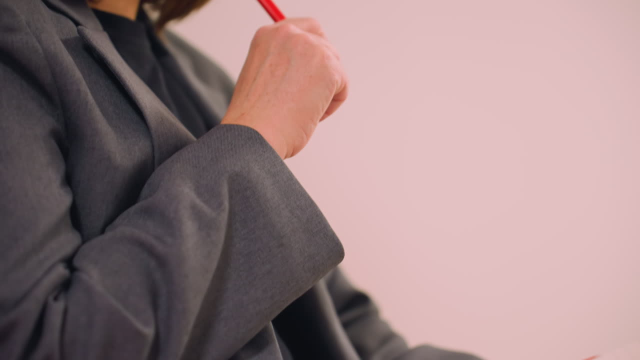Close-up of businesswoman in dark blazer writing in notebook with red pen, showing hand movement and part of body in focused work environment, representing productivity, planning, and office activity