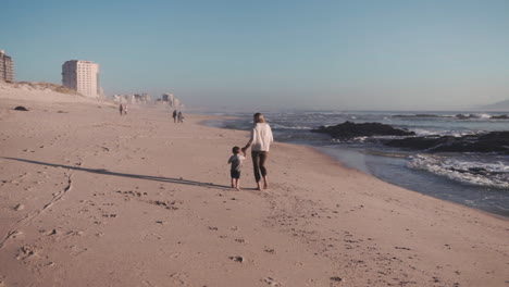 Mother-and-son-walking-barefoot-in-the-beach-and-bonding-together-during-family-vacations,-back-view