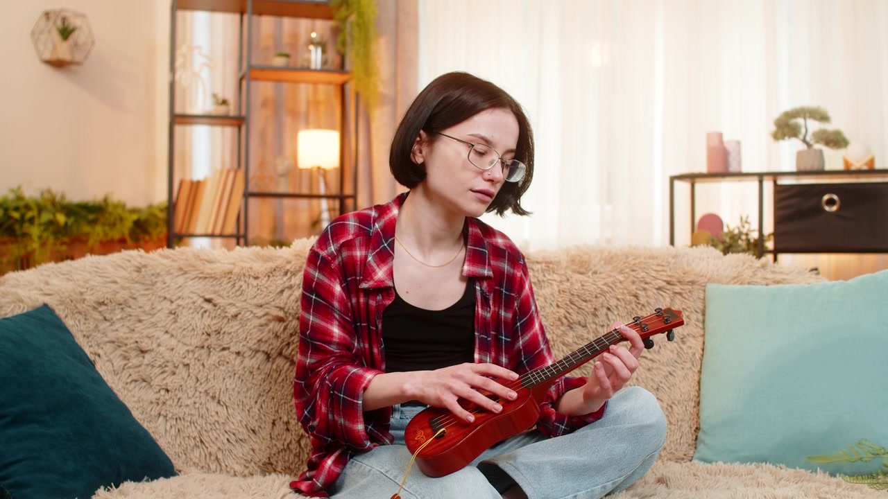 Wanita muda bermain ukulele di rumah fokus mempelajari akord dan tersenyum dengan kepuasan dan kegembiraan