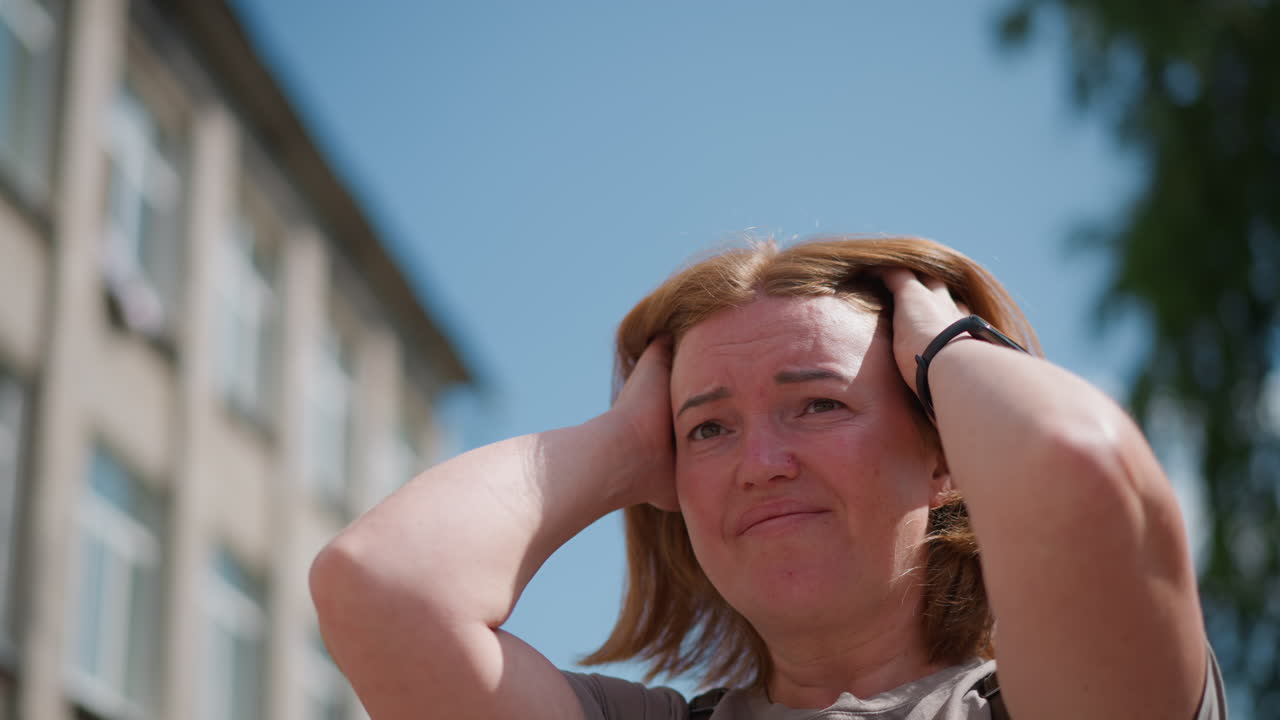 Sad pupil stands outdoors near residential area, adjusting hair with hand wearing smart watch, subtle facial tension under sunlight expresses loneliness, worry, and thoughtful emotion