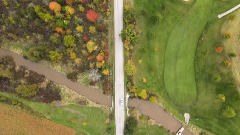 car driving moving along straight rural countryside road in autumn season, path through green fields lands trees and country homes, aerial top down tracking view