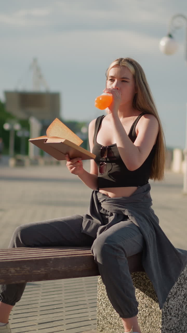 mujer sentada al aire libre sosteniendo un libro en una mano mientras beba jugo de la botella con la otra, centrada en la lectura, el fondo presenta postes de lámparas y vegetación