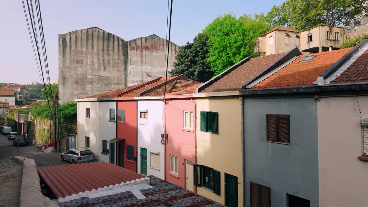 Vista panoramica di una vivace fila di case tradizionali di Porto, ognuna dipinta con un colore diverso e con classici tetti in tegole. Cavi elettrici attraversano il cielo e un vecchio edificio industriale si erge sullo sfondo.