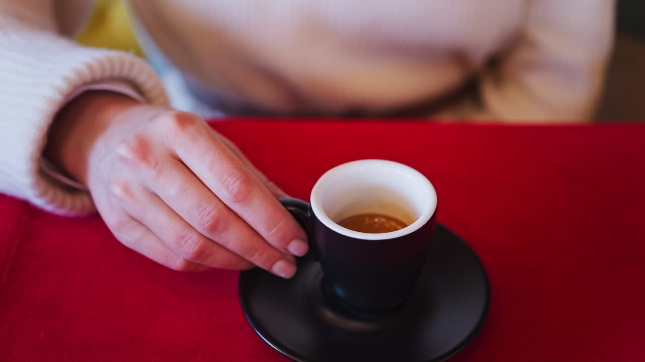 Close up of a woman holding an espresso in a black cup on a red tablecloth at a restaurant