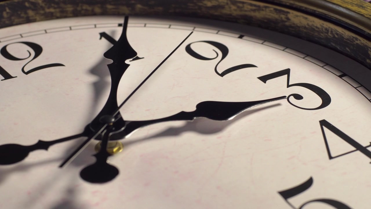 Primo piano di un quadrante di orologio analogico vintage. La lancetta dei secondi ticchetta in senso orario sui numeri romani mentre la telecamera si muove in una lenta panoramica verso destra. Scatto macro sotto luce da studio interna.
