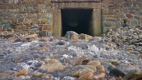 Cascada-De-Agua-Desde-Un-Agujero-En-Un-Muro-De-Piedra-A-Través-De-Una-Playa-Pedregosa-En-Broad-Haven,-Gales-Occidental