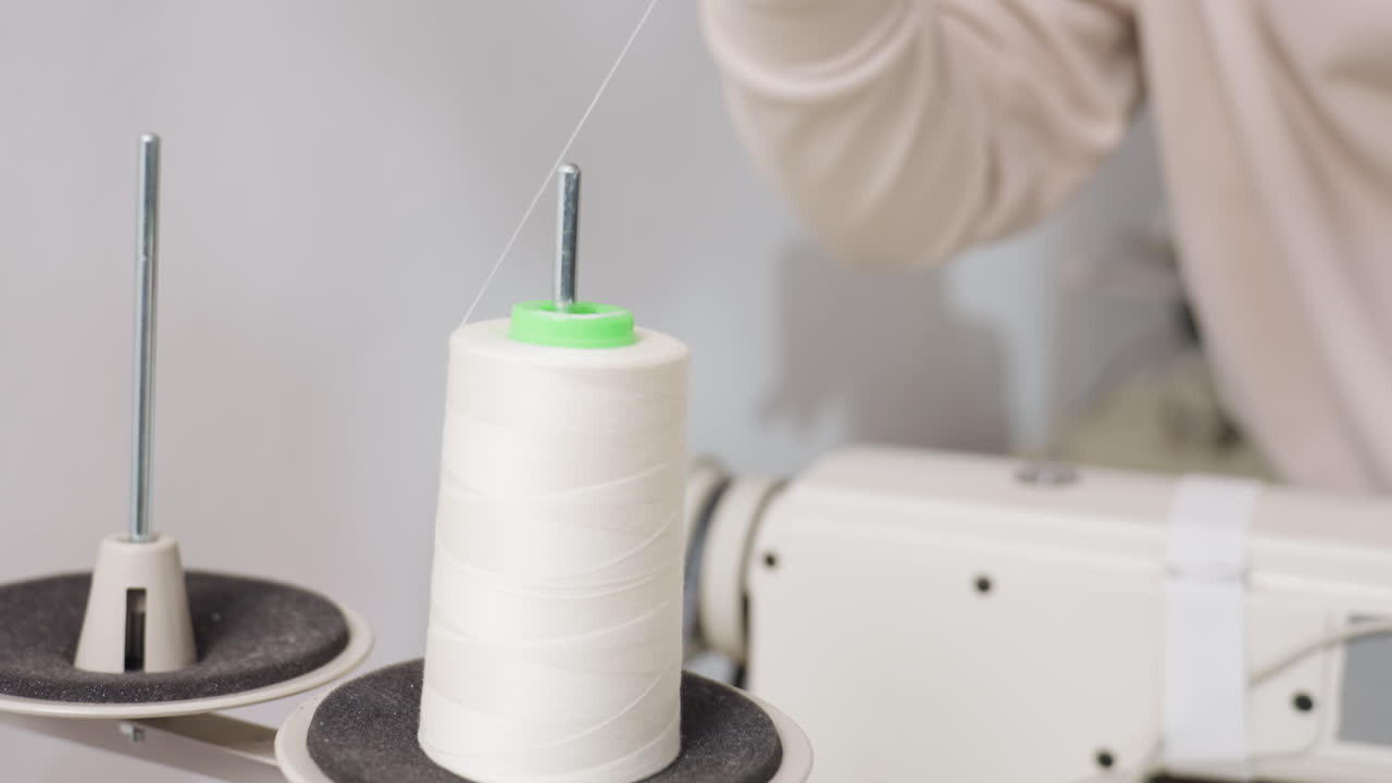 Dress maker carefully positioning big white spool onto thread guide of sewing machine, showcasing concentration, dexterity, and professional skill in textile workspace, focusing on precise technique