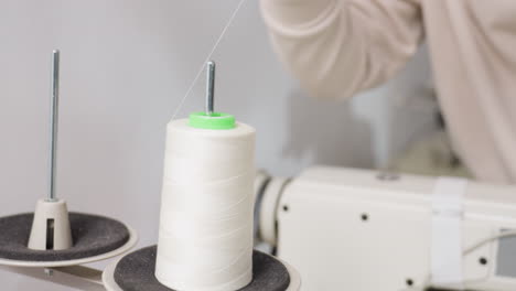 Dress maker carefully positioning big white spool onto thread guide of sewing machine, showcasing concentration, dexterity, and professional skill in textile workspace, focusing on precise technique