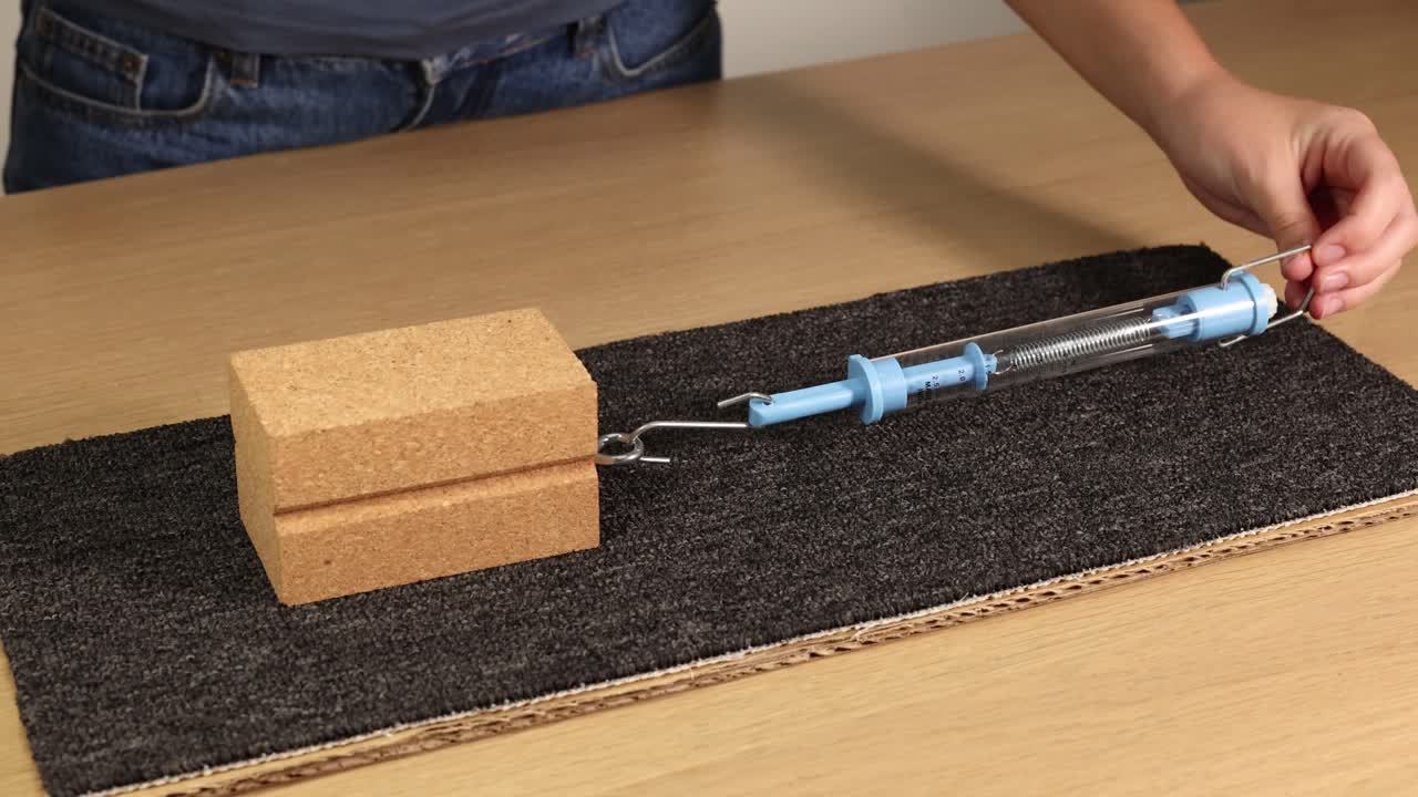 A hand uses a spring scale to measure friction force on a wooden block over a carpet surface, highlighting physics principles