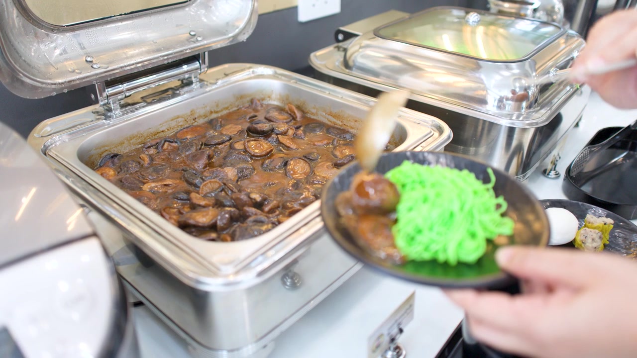 Un cameriere usa un mestolo per servire uno stufato di funghi da uno scaldavivande in acciaio inossidabile.
