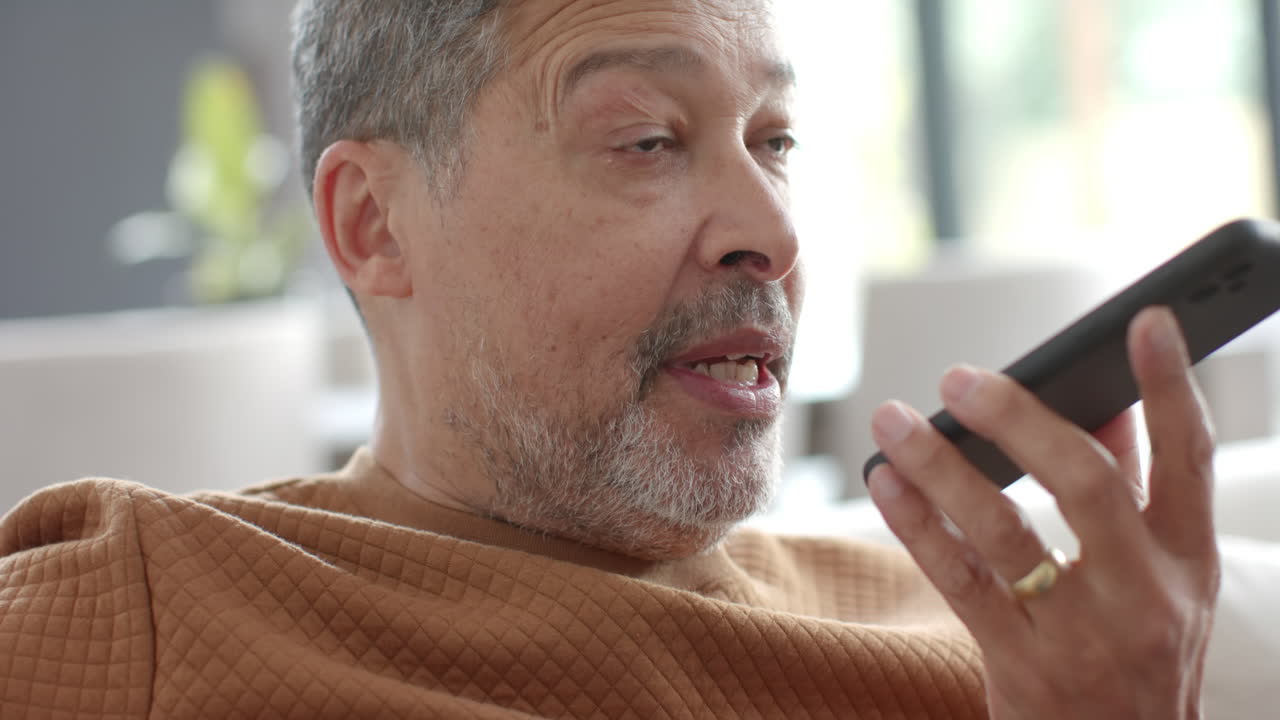Senior biracial man talking on smartphone sitting in sunny living room, slow motion