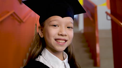 zoom in of a happy preschool female student in cap and gown holding graduation diploma and looking at the camera