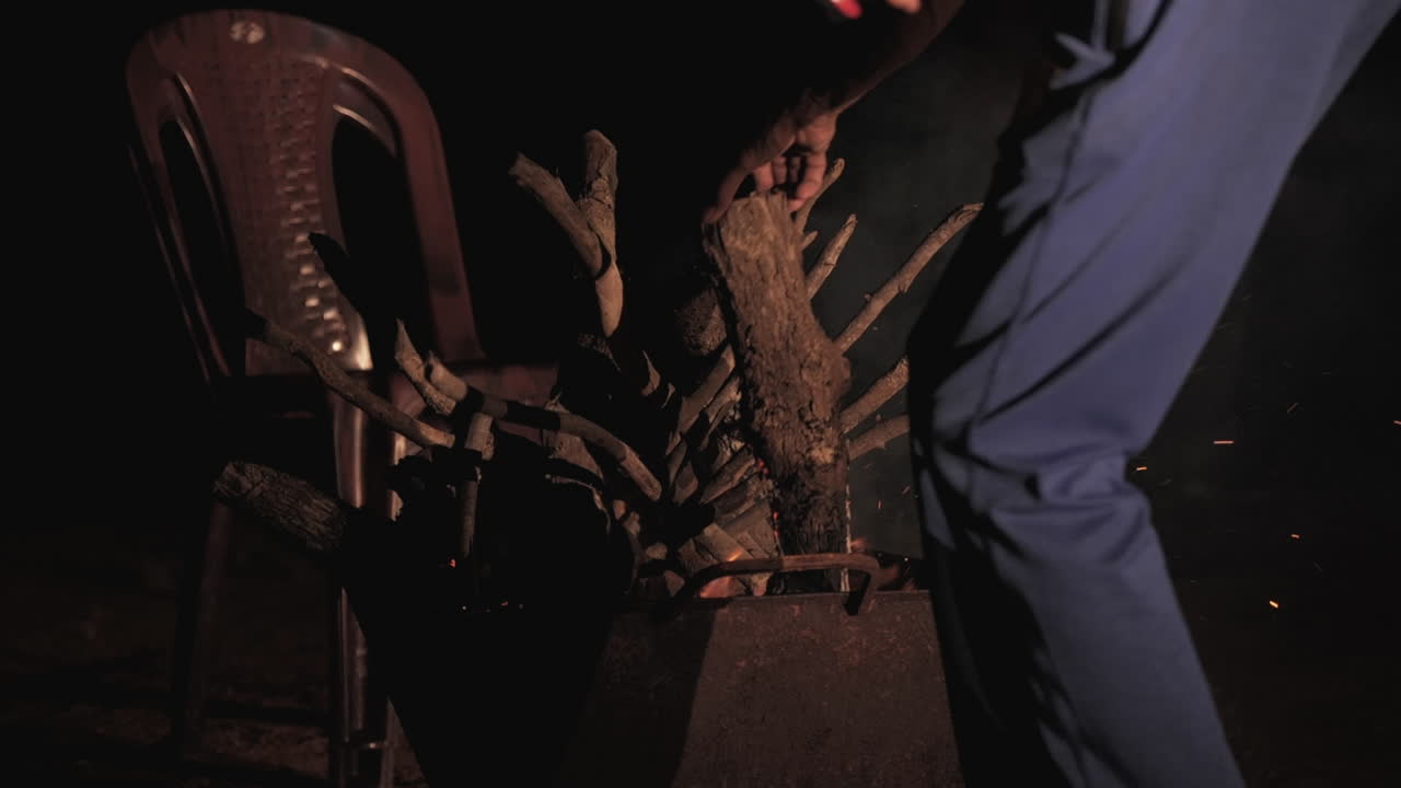 A bonfire burns brightly at night, with logs and chairs around in a slow-motion scene